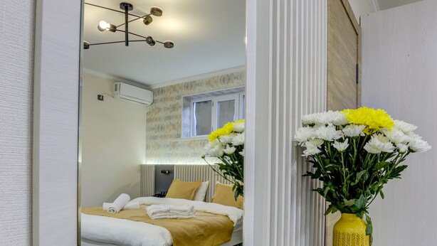 Une chambre à coucher moderne avec un grand miroir, un lit avec une literie jaune et un vase rempli de fleurs posé sur une console.
