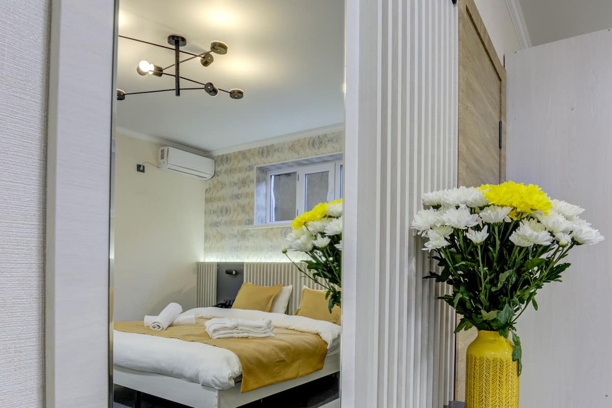 Un dormitorio moderno con un espejo grande, una cama con ropa de cama amarilla y un florero con flores sobre una consola.