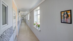 Un couloir aux murs blancs, doté d’un sol carrelé et agrémenté de plantes en pot posées sur les rebords des fenêtres.