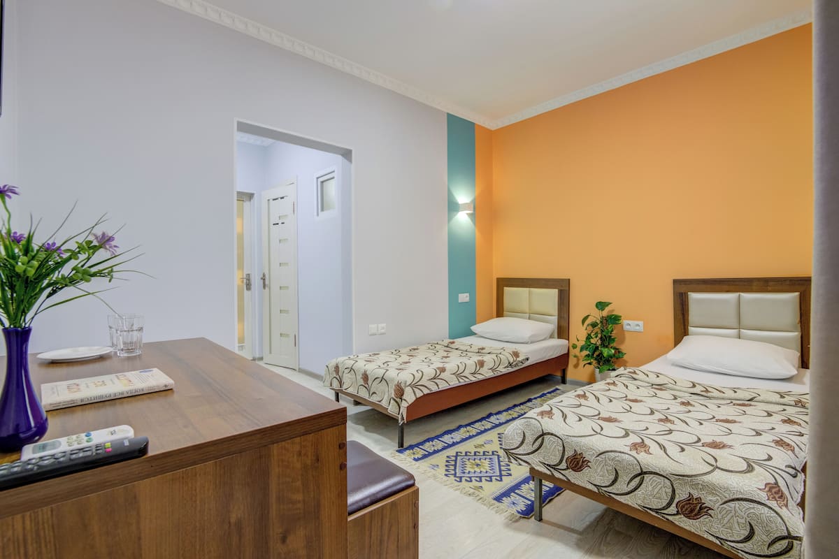 Habitación de hotel con dos camas, un escritorio con un florero, una mesita con un control remoto y una puerta que da a otra habitación.