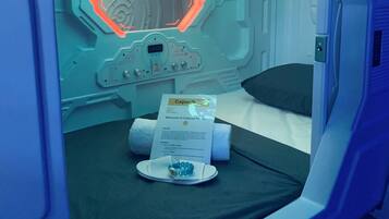 Camera in hotel a capsule con un letto, una sveglia digitale, un cartello di benvenuto e un piccolo vassoio con degli oggetti.