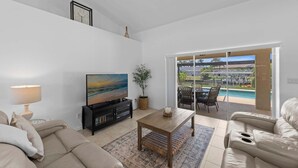 Sala de estar con televisor, sofá, mesa de centro y vista a una piscina a través de puertas corredizas de vidrio.