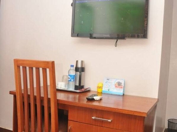 Un escritorio de madera con un televisor montado encima, una silla, una botella de agua, una cafetera, un control remoto y una tarjeta pequeña.