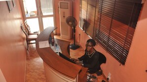A reception area with a wooden desk, a fan, and a window with blinds. - Eeyilobe Guest House (Lagos)