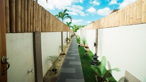 A narrow pathway flanked by wooden fences and white walls, leading to a small garden area with potted plants and a decorative stone feature.