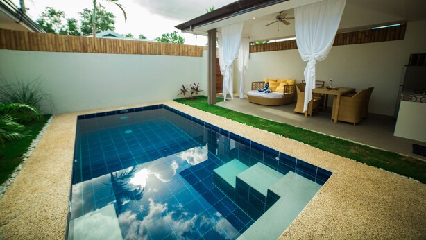 A swimming pool with a tiled deck, a covered seating area with cushions, and a wooden fence.