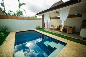 Une piscine avec une terrasse carrelée, un espace détente couvert avec des coussins et une clôture en bois.