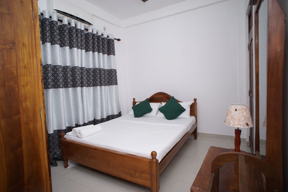 Una cama bien hecha con estructura de madera, una mesita de noche con lámpara y una cortina estampada.