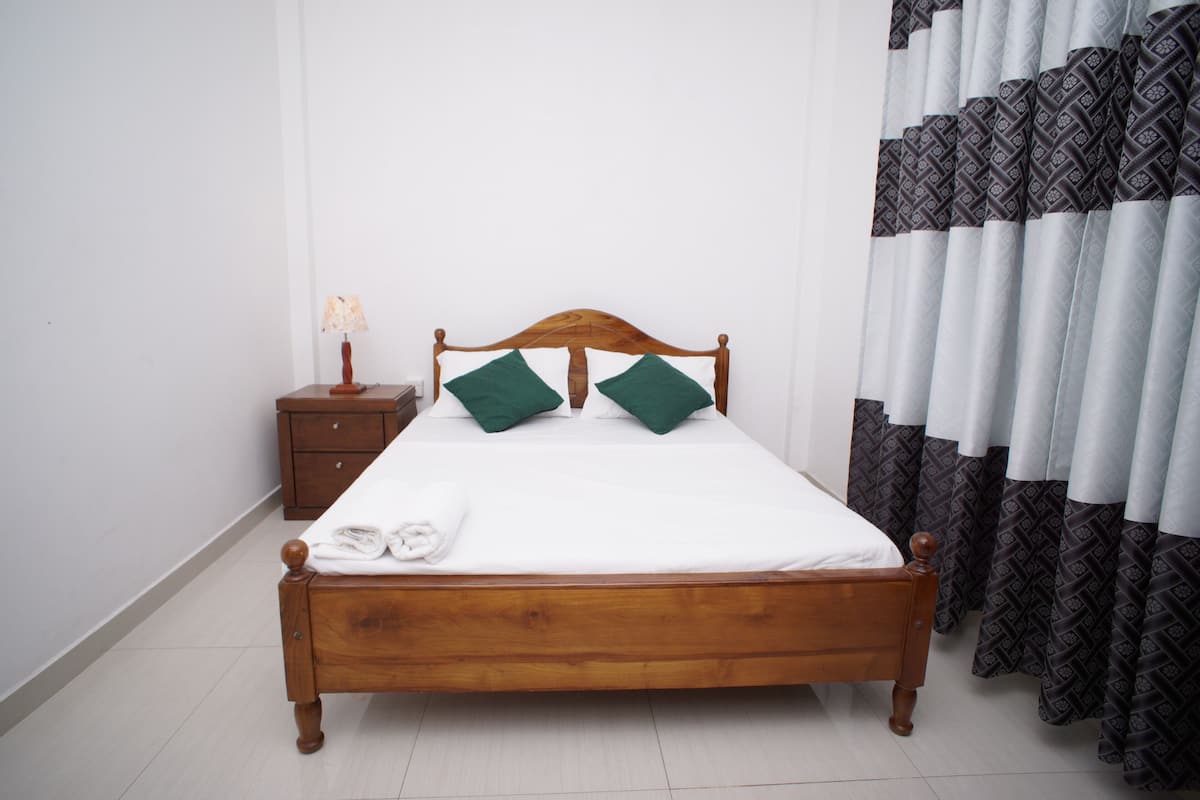 Una cama bien tendida con cabecera de madera, una mesita de noche con lámpara y cortinas estampadas.