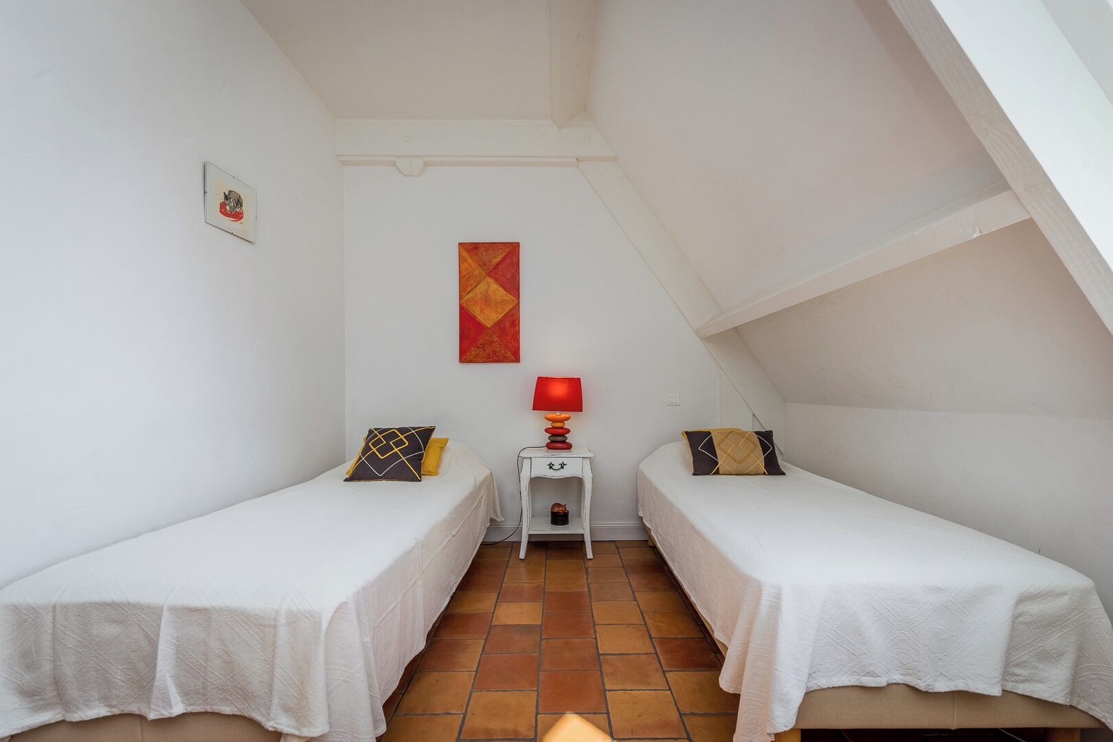 A small, white-walled room with two single beds, a small table with a red lamp, and a red and yellow abstract painting on the wall.