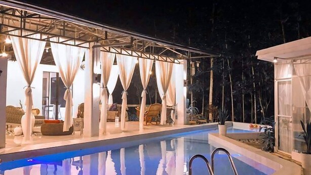 An outdoor swimming pool with a wooden deck, surrounded by a covered seating area with white curtains. - Balconahe Resto Camp (Antipolo)