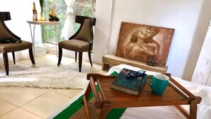A room with a bed, a wooden side table with a book and a mug, two chairs, a painting, and a view of greenery through the window. - Balconahe Resto Camp (Antipolo)