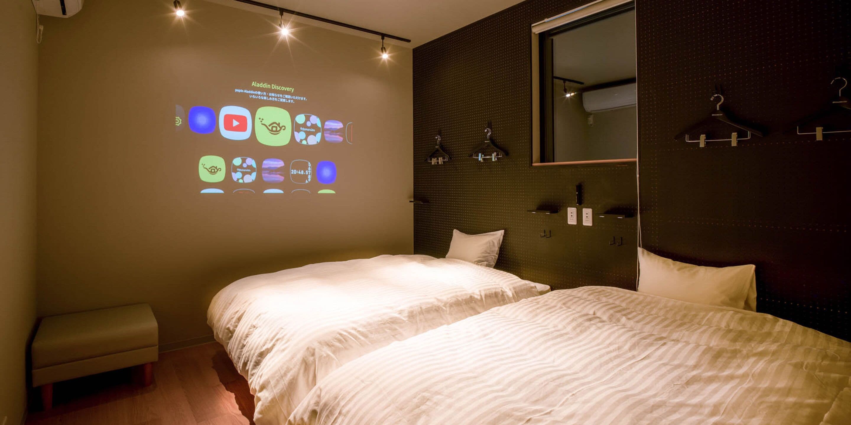 Une chambre d’hôtel avec deux lits, un projecteur fixé au mur diffusant une présentation, un miroir et une armoire avec des cintres.