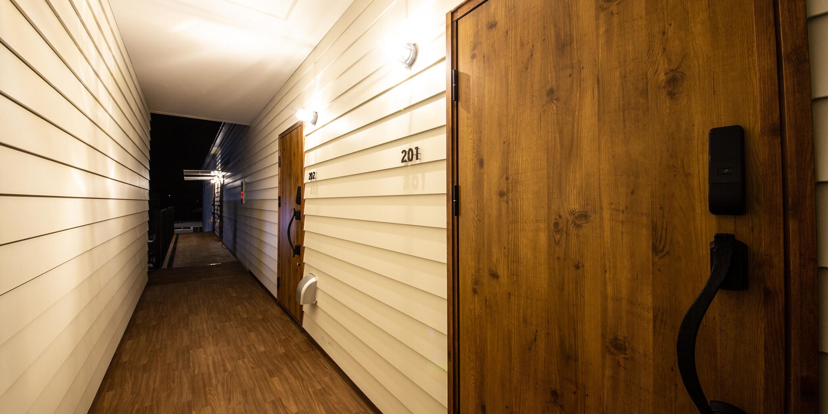 A narrow hallway with wooden floors and white walls, featuring a wooden door with a number plate and a black handle.