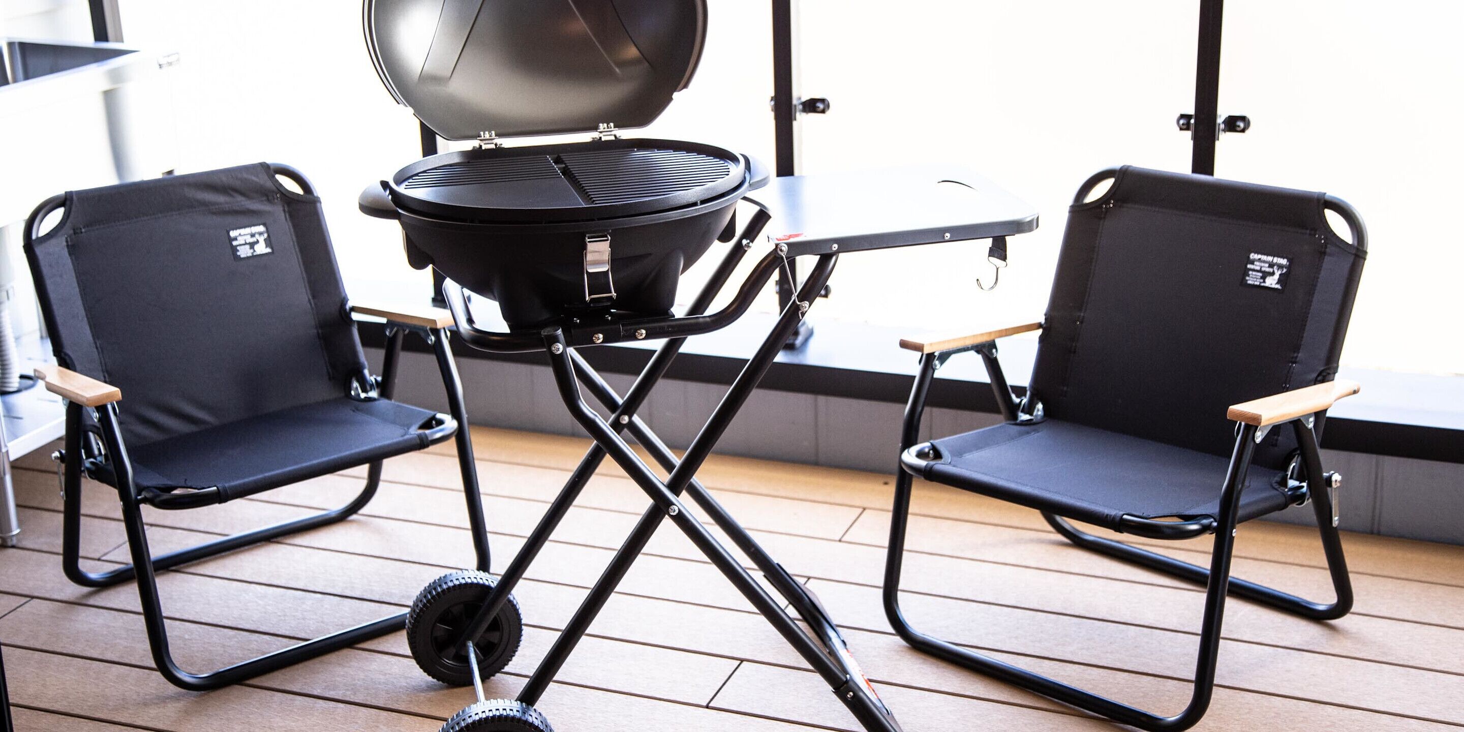 Deux chaises pliantes noires avec des accoudoirs en bois, un barbecue portable et une table en métal, le tout placé sur une terrasse en bois.
