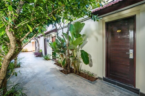 A corridor with a tiled floor, a wooden door with a number plate, and various plants along the wall. - Kelantel Bali (Tuban)