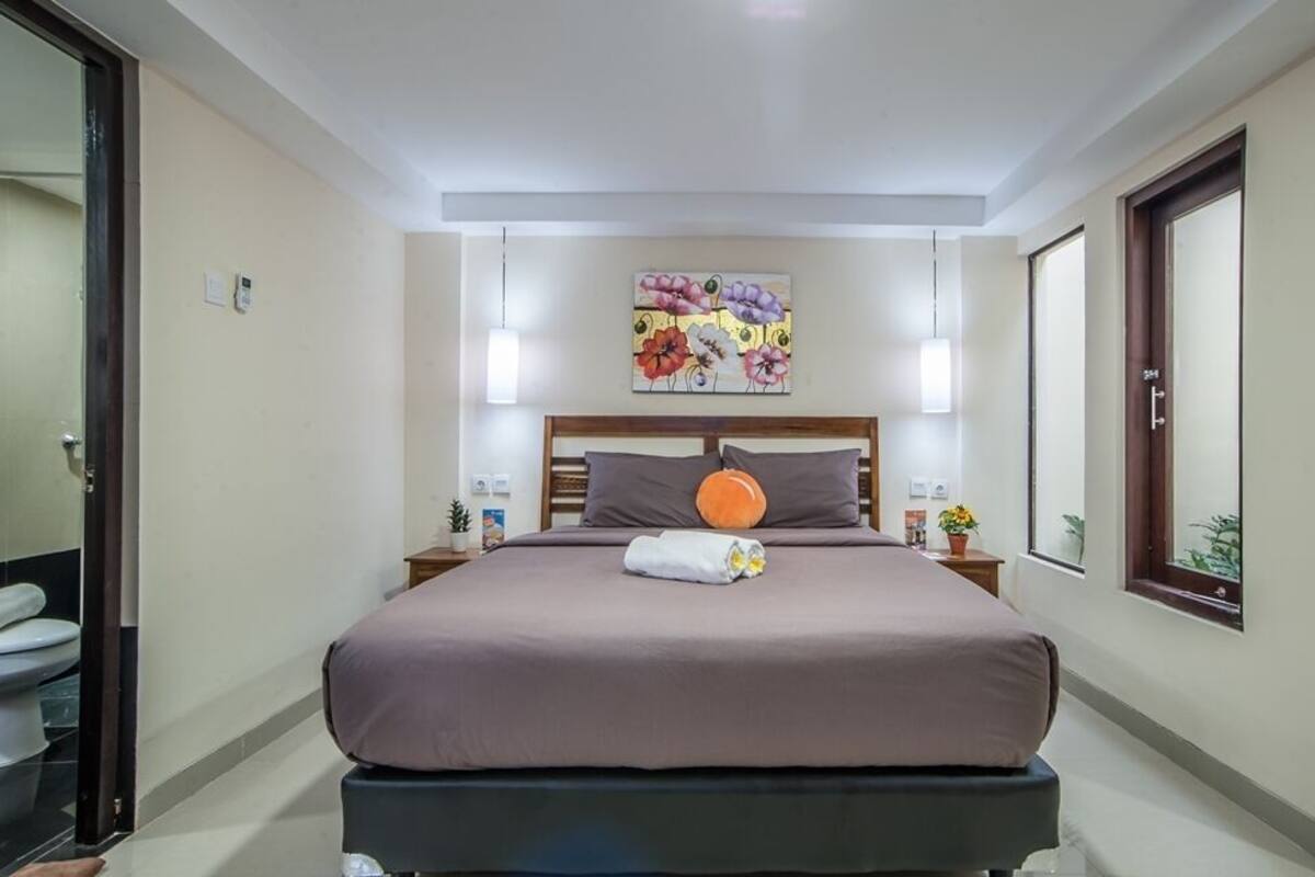 Ett hotellrum med en stor säng, en sänggavel med ett blomstermotiv, sänglampor, ett litet bord med en växt och ett fönster med utsikt.