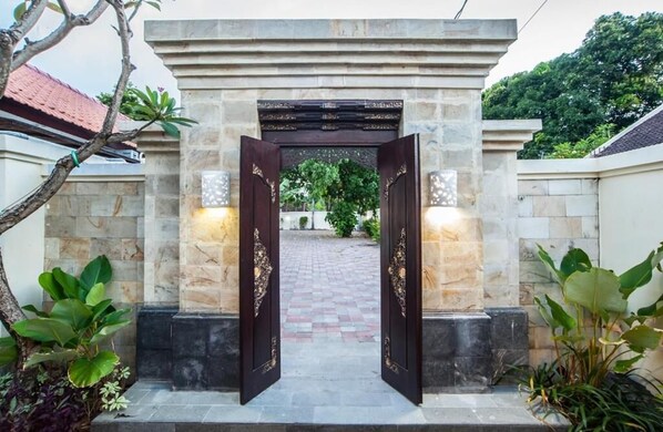 A stone entrance with a dark wooden door, flanked by two lantern-style lights, leading to a paved pathway. - Kelantel Bali (Tuban)