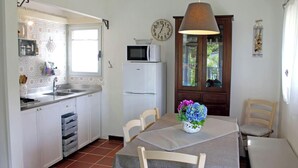 A kitchen with white cabinets, a sink, a microwave, and a refrigerator. There is a dining table with chairs and a vase of flowers. - Scaglieri Mare - ibookingelba.com (Portoferraio)