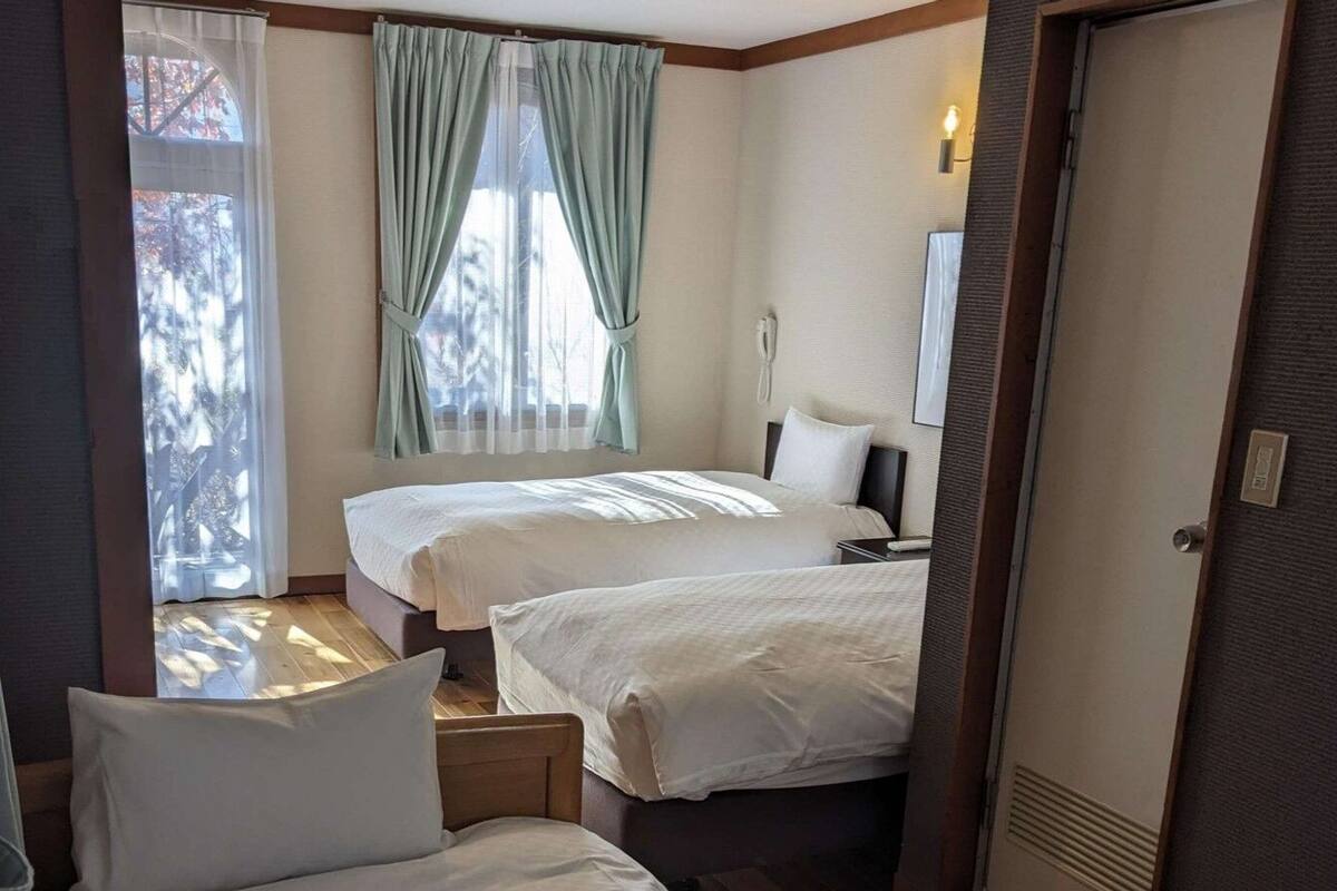 A hotel room with two beds, a window with curtains, a bedside table, a lamp, and a door.