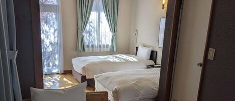 Habitación de hotel con dos camas, una ventana con cortinas, una mesita de noche, una lámpara y una puerta.