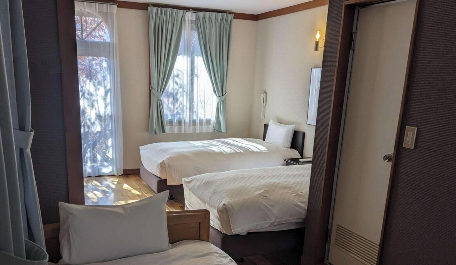 A hotel room with two beds, a desk, a chair, a window with curtains, a bedside table, and a door.