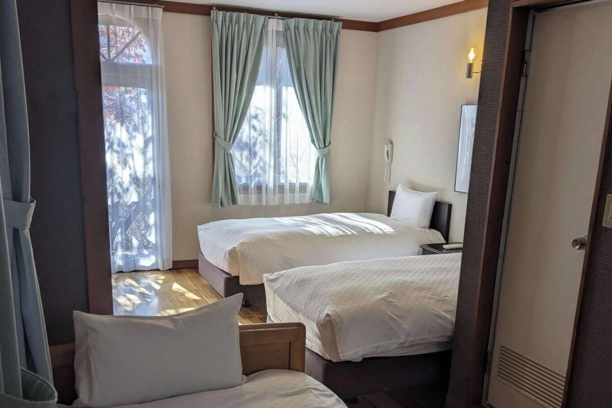 A hotel room with two beds, a desk, a chair, a window with curtains, a bedside table, and a door.
