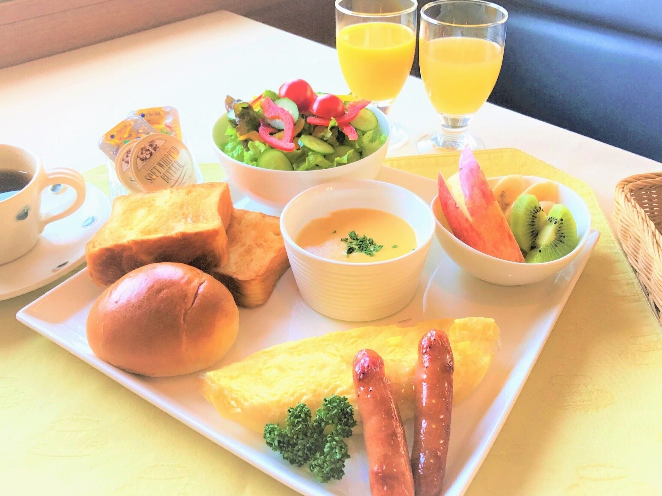 A breakfast plate with toast, sausages, scrambled eggs, a bowl of soup, a salad, and a bowl of fruit.