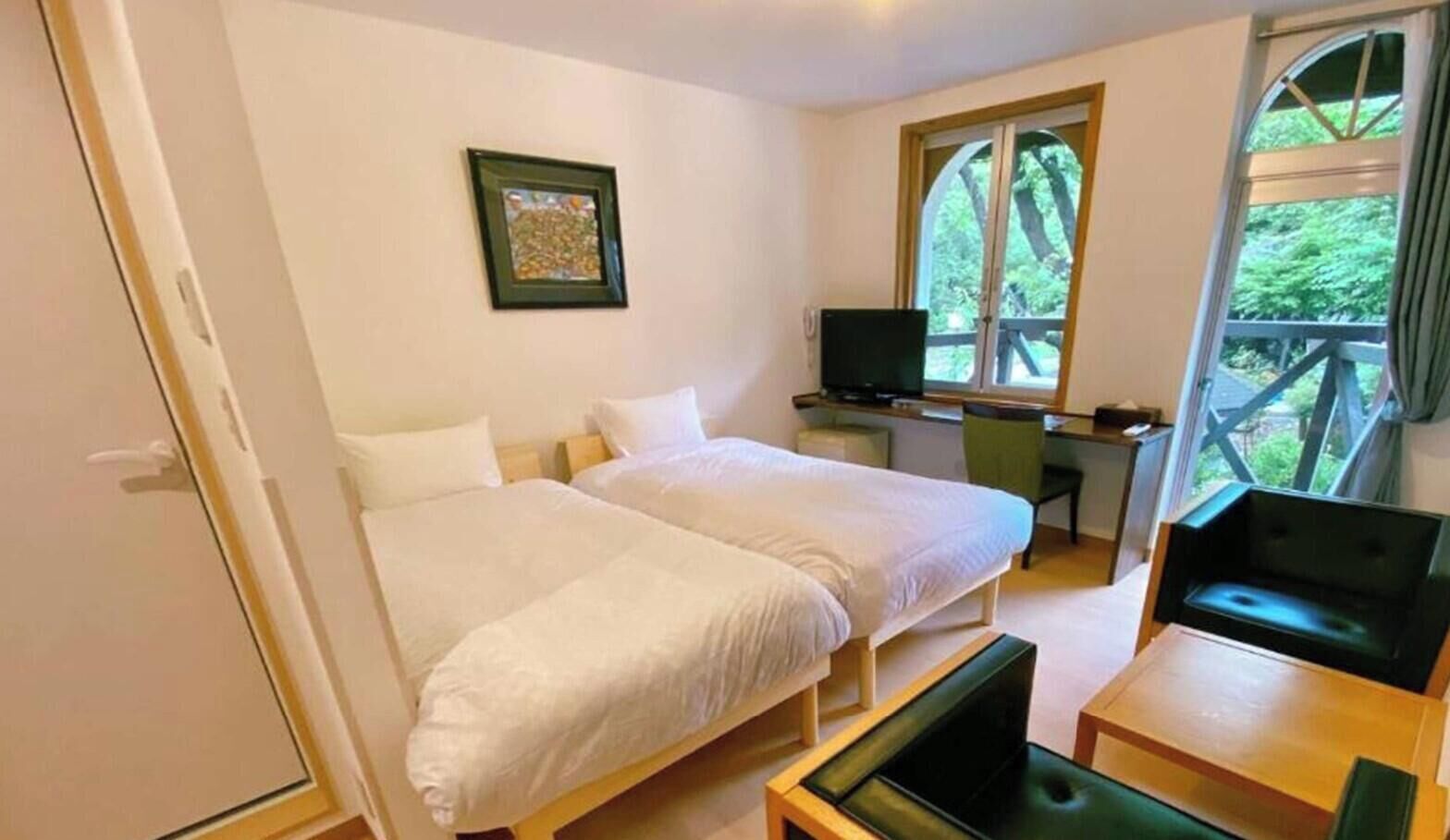 A room with two beds, a desk, a chair, a TV, and a window with a view of trees.
