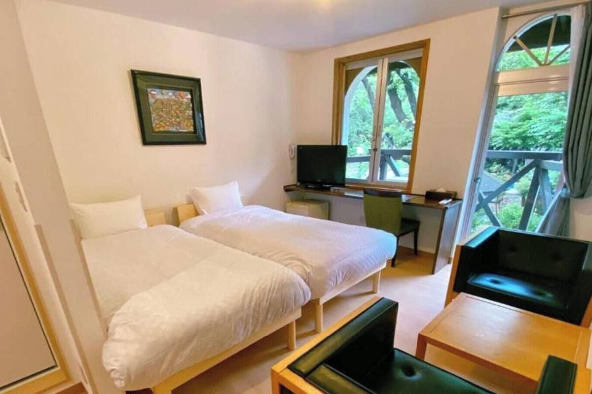 A room with two beds, a desk, a chair, a TV, and a window with a view of trees.