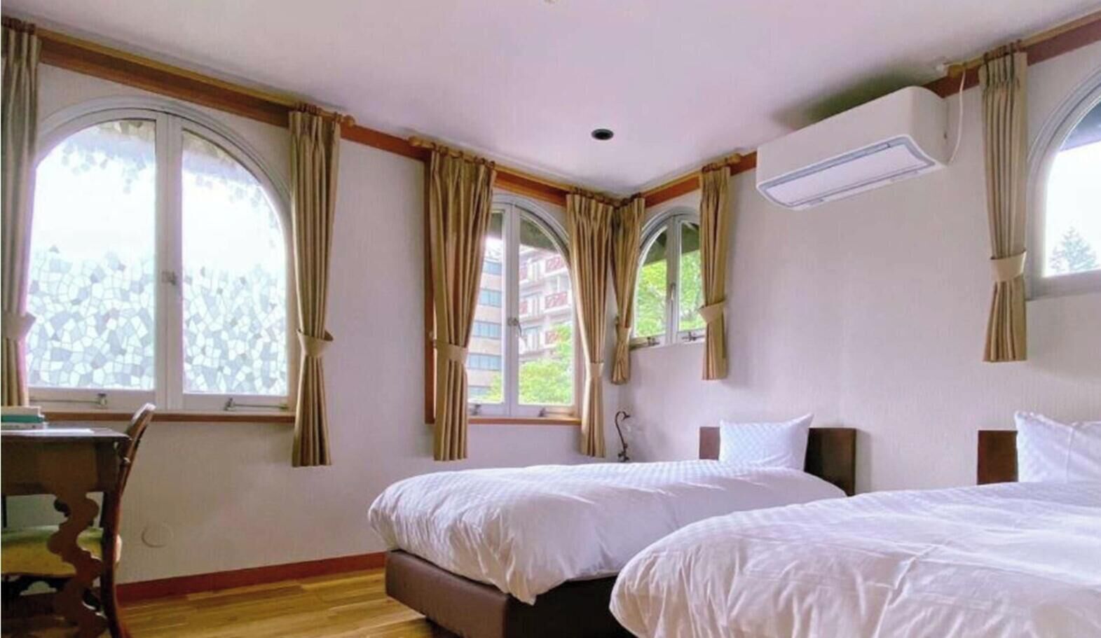 A hotel room with two beds, a desk, and large windows with curtains.
