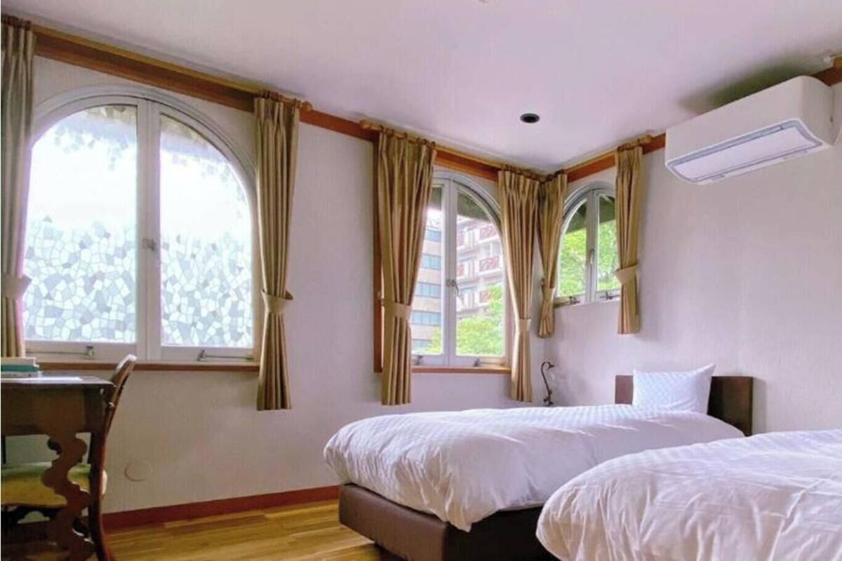 A hotel room with two beds, a desk, and large windows with curtains.