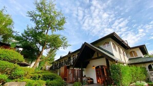 A two-story building with a balcony, surrounded by greenery and a gravel driveway. - Lodge Atelier (Hokuto)