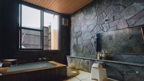 A bathroom with a stone wall, a bathtub, a window, and a shelf with toiletries. - Lodge Atelier (Hokuto)