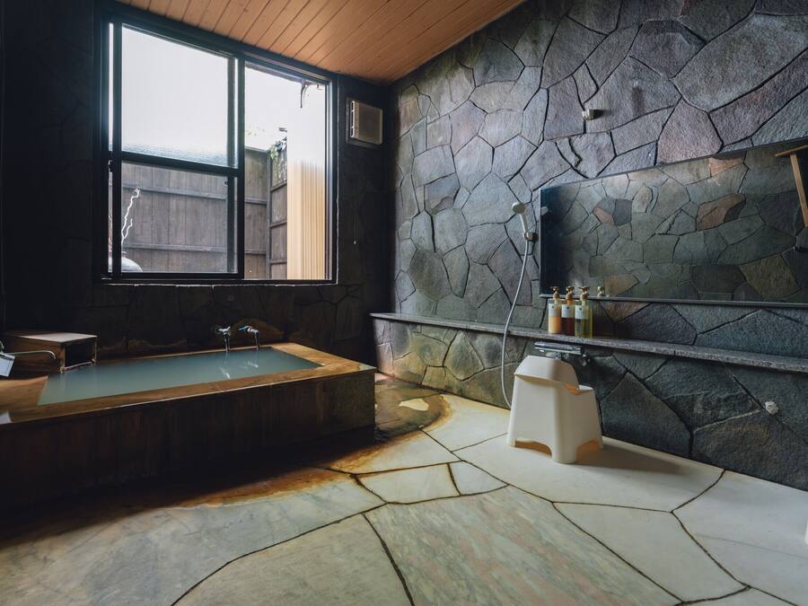 A bathroom with a stone wall, a bathtub, a window, and a shelf with toiletries.