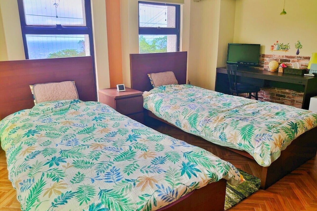 A room with two beds, each with a floral-patterned bedspread, a small bedside table, a TV, and a brick accent wall.