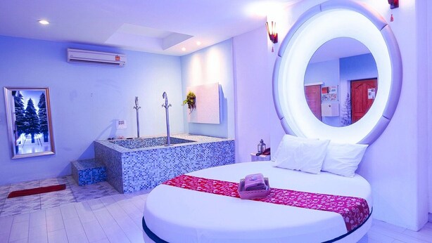 Un dormitorio moderno con cama redonda, un espejo circular empotrado, un pequeño rincón de lectura con una silla y una mesa, y un amplio ventanal con vistas a árboles.
