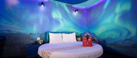 A circular bed with a white mattress and floral bedspread, a blue headboard, and two red slippers on the side.