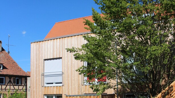 Un edificio moderno con fachada de madera, techo rojo y un pequeño jardín con un árbol y arbustos.