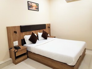 A neatly made bed with white linens and brown pillows, a wooden headboard, a bedside table with a phone, and a wall-mounted picture above the bed. - Hexa Royal Elite (Mumbai)