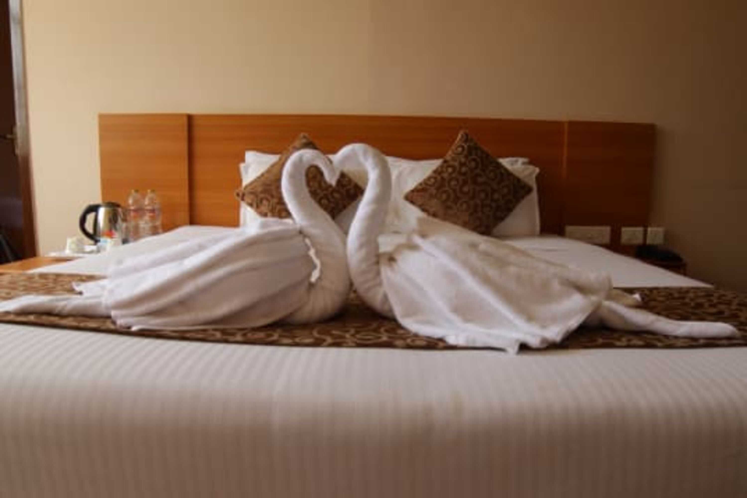 Una cama bien hecha con cabecera de madera, un cojín decorativo y un par de toallas dispuestas en forma de corazón.