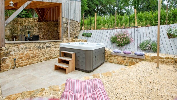 A hot tub in a stone-walled outdoor area with steps leading up to it, surrounded by a gravel path and a wooden deck. - Dreamy Retreat in Loubejac (Loubejac)