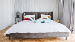 Un dormitorio con una cama grande, cortinas grises, una mesita de noche pequeña con una planta y una piña sobre la mesita.