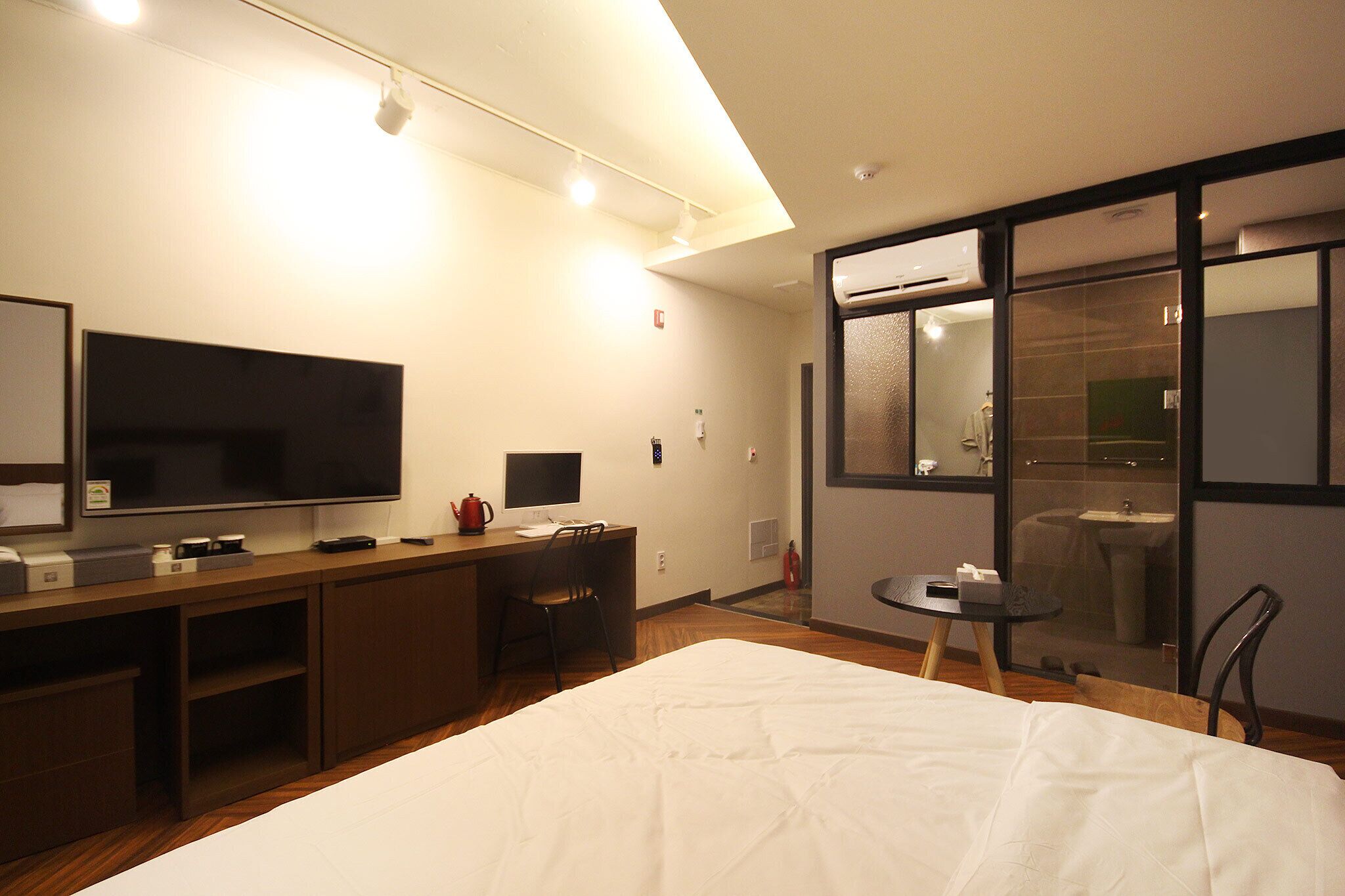 A hotel room with a large bed, a desk with a flat-screen TV, a small table, and a bathroom visible through a glass door.