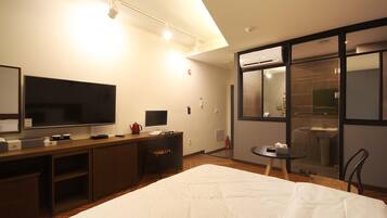 A hotel room with a large bed, a desk with a flat-screen TV, a small table, and a bathroom visible through a glass door.