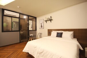 A hotel room with a large bed, a wooden floor, a glass shower enclosure, a small table, and a chair.