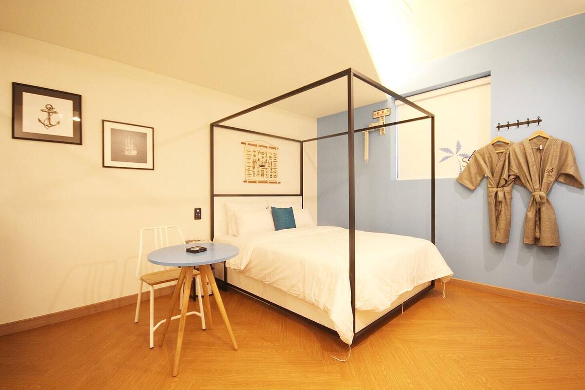 A bedroom with a four-poster bed, a small round table with a chair, and a wall-mounted robe rack.