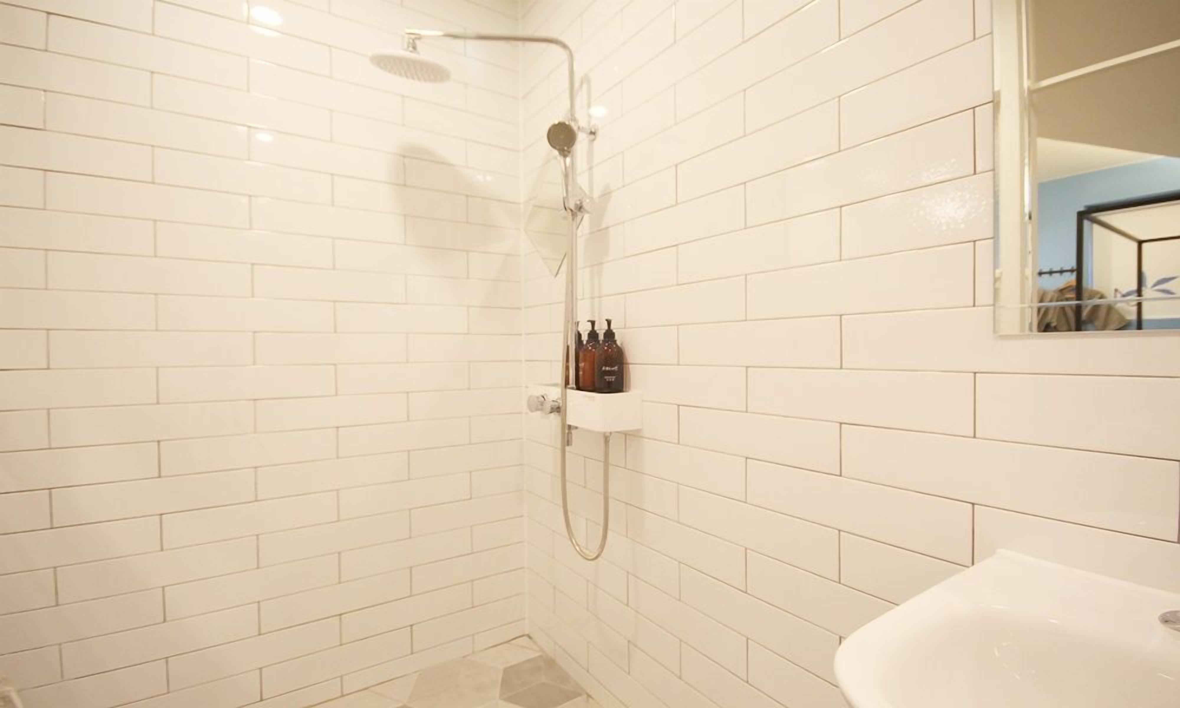 Une salle de bain avec des murs recouverts de carreaux blancs, une douche, un lavabo et un miroir.
