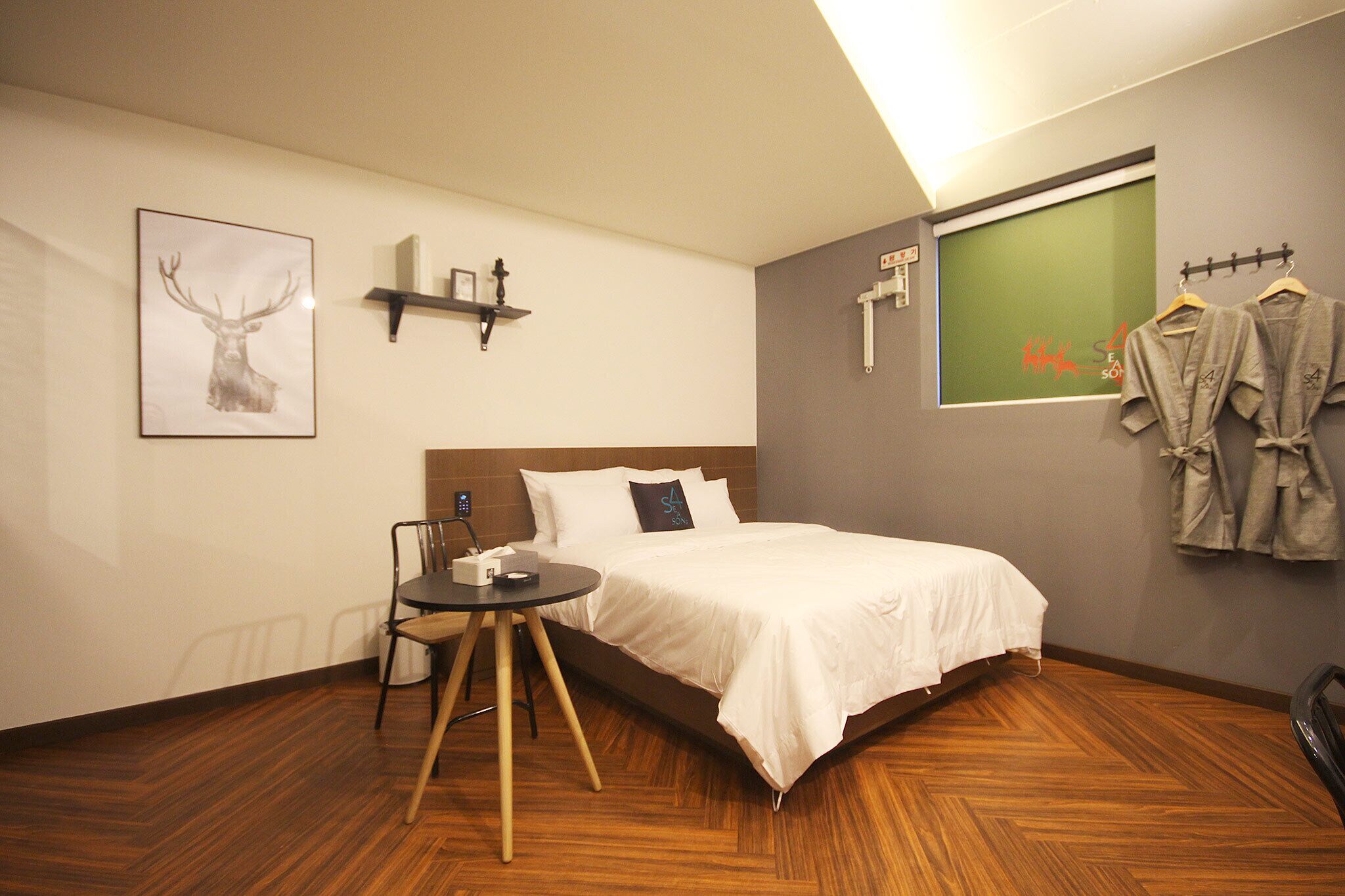 A hotel room with a bed, a bedside table with a lamp, a chair, a picture of a deer, a coat rack with jackets, and a window with a green view.
