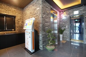 A reception area with a self-service kiosk, a potted bonsai tree, and a glass door leading to the elevator.